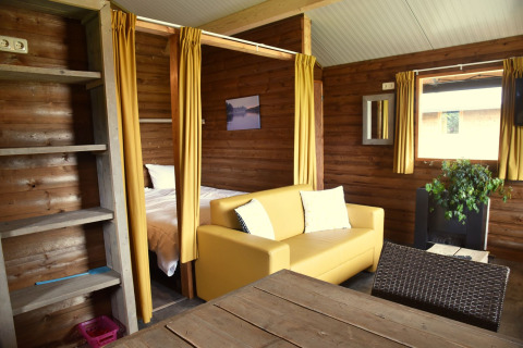 Interior acogedor de cabaña en Camping de Koeksebelt, Países Bajos, con sofá amarillo y paredes de madera.