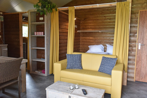 Interno di cabina in legno con divano giallo e tende al Camping de Koeksebelt nei Paesi Bassi.