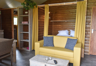 Interno di cabina in legno con divano giallo e tende al Camping de Koeksebelt nei Paesi Bassi.