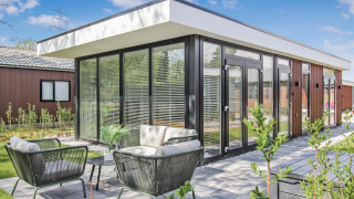 Moderne Forest villa lodge met grote ramen, terras en tuinmeubilair onder een heldere blauwe lucht.