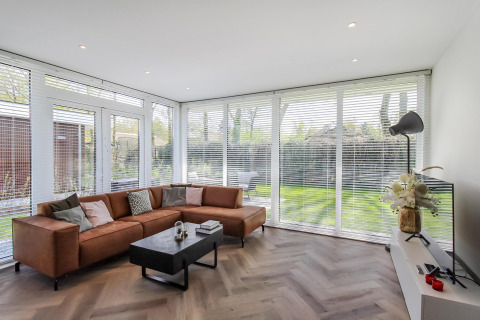 Stilfuld stue i Forest villa lodge med store vinduer, brun sofa og havedesign synligt udenfor.