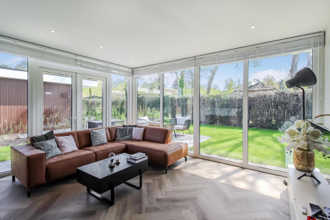 Moderne stue i Forest villa-lodge med brune lædersofaer, store vinduer og udsigt til haven.