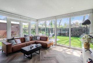 Moderne stue i Forest villa-lodge med brune lædersofaer, store vinduer og udsigt til haven.