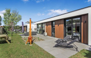 Moderne Forest villa ved Uniq Leisure, Holland, med loungeområde, liggestole og grøn have under blå himmel.