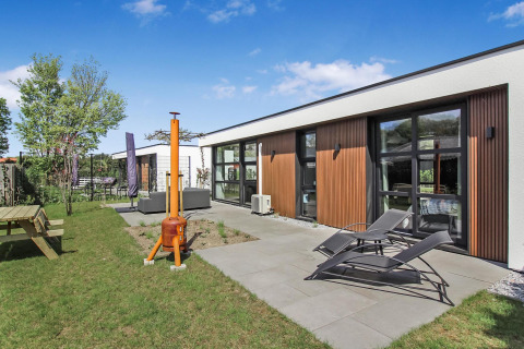 Moderne Forest villa ved Uniq Leisure, Holland, med loungeområde, liggestole og grøn have under blå himmel.