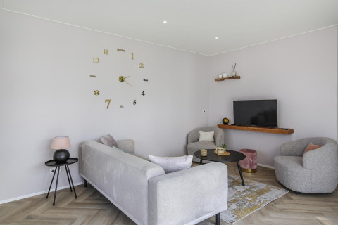 Sala de estar moderna con muebles grises, reloj de pared y TV en Eco Lodge de Uniq Leisure, Países Bajos.