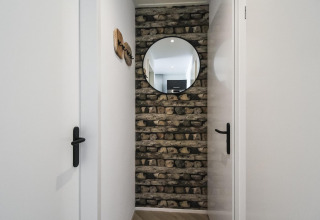 Pasillo estrecho con pared de piedra, espejo redondo y puertas blancas en Eco Lodge en Uniq Leisure, Países Bajos.