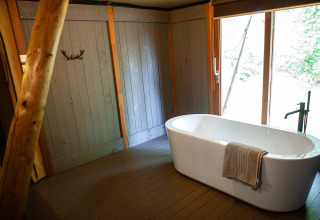 Bathroom in a safari tent with a freestanding bathtub, wood panel walls, and large windows at Kings Lodge, Sandberghe.