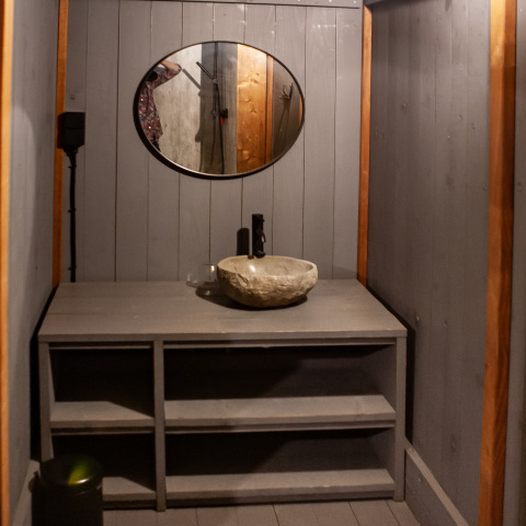 Baño rústico con lavabo de piedra, estanterías grises y espejo redondo en Kings Lodge, Sandberghe, Países Bajos.