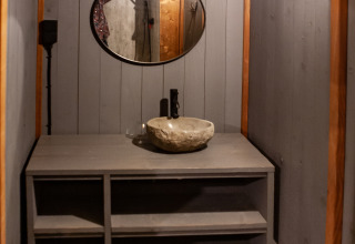Baño rústico con lavabo de piedra, estanterías grises y espejo redondo en Kings Lodge, Sandberghe, Países Bajos.