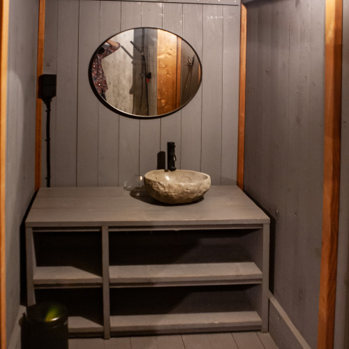 Rustic bathroom with stone sink, grey wooden shelves and round mirror in Kings Lodge, Sandberghe, Netherlands.