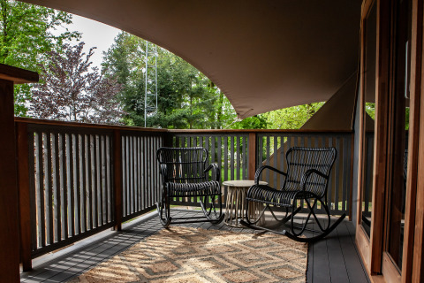 Terraza cubierta con dos mecedoras y mesa en Kings Lodge + Hot Tub, Sandberghe, Países Bajos, rodeada de árboles.