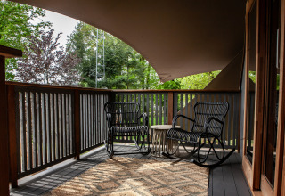 Terraza cubierta con dos mecedoras y mesa en Kings Lodge + Hot Tub, Sandberghe, Países Bajos, rodeada de árboles.