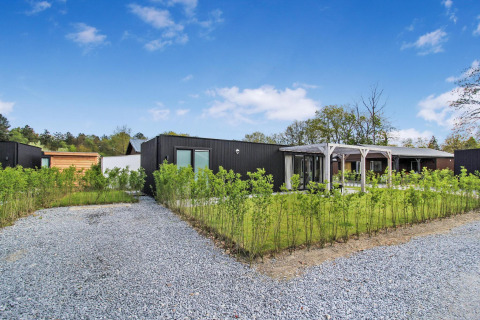 Moderne Eco Lodge med sort facade, grusvej, grønne planter og blå himmel på en solrig dag.