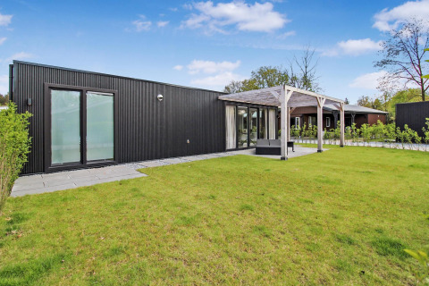 Moderne Eco Lodge med sort facade, store vinduer og terrasse, omgivet af grøn græsplæne og blå himmel.