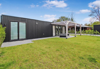 Moderne Eco Lodge med sort facade, store vinduer og terrasse, omgivet af grøn græsplæne og blå himmel.