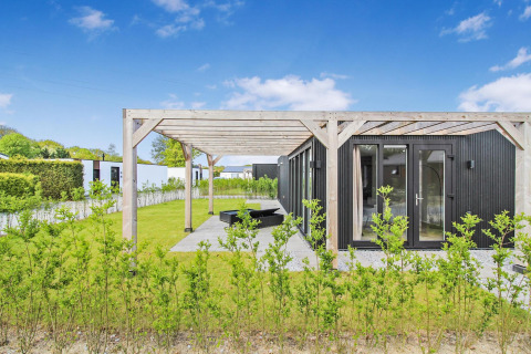 Eco Lodge moderna de fachada negra con pérgola de madera y jardín verde bajo un cielo azul claro.