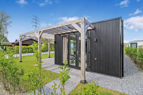 Eco Lodge negra moderna con pérgola de madera, grandes ventanales y jardín verde bajo cielo azul claro.