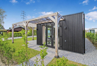 Eco Lodge negra moderna con pérgola de madera, grandes ventanales y jardín verde bajo cielo azul claro.