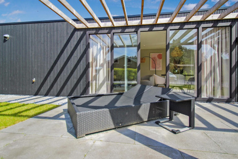 Terraza soleada en Eco Lodge con fachada negra moderna, tumbona de mimbre y grandes ventanales al salón.