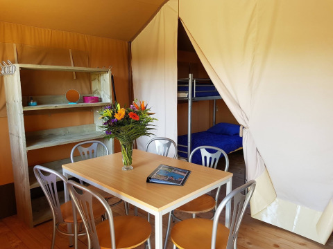 Inde i en safaritelts stue med et bord, stole, blomster, reol og udsigt til køjesenge på De Scherpenhof.