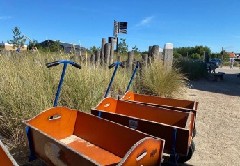 Houten bolderkarren langs een zandpad bij hoog gras op een zonnig glampingterrein, klaar om te trekken.