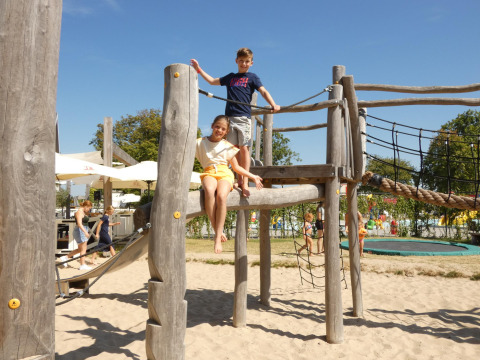 Niños juegan en un parque infantil de madera sobre arena en un alojamiento glamping, familias al fondo.