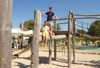 Niños juegan en un parque infantil de madera sobre arena en un alojamiento glamping, familias al fondo.