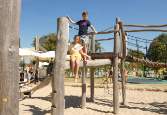 Niños juegan en un parque infantil de madera sobre arena en un alojamiento glamping, familias al fondo.
