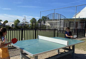 Two kids play outdoor table tennis at a glamping site, with tents and facilities visible in the background.