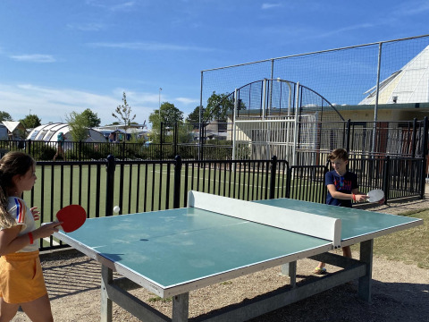 Two kids play outdoor table tennis at a glamping site, with tents and facilities visible in the background.