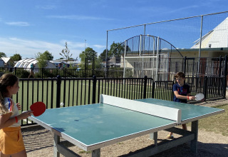 Dos niños juegan al ping-pong al aire libre en un alojamiento glamping, con tiendas y áreas visibles detrás.