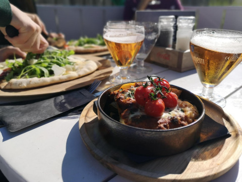 Comida al aire libre en glamping: pizza con rúcula, plato horneado y cerveza en mesa soleada.