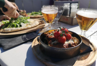 Comida al aire libre en glamping: pizza con rúcula, plato horneado y cerveza en mesa soleada.