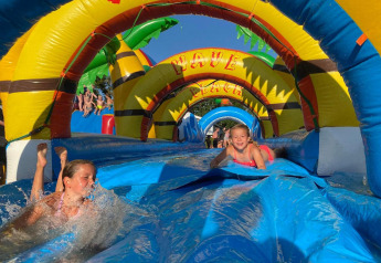 Niños disfrutan deslizándose por un tobogán inflable de agua en un sitio de glamping con arcos coloridos.
