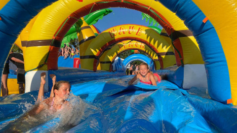 Niños disfrutan deslizándose por un tobogán inflable de agua en un sitio de glamping con arcos coloridos.