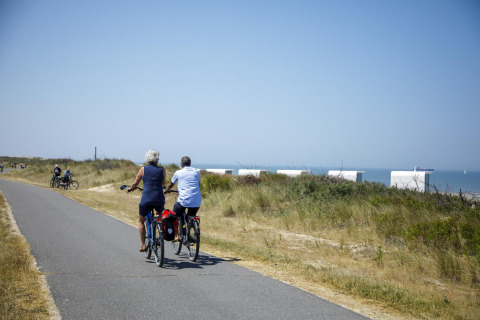 Zwei Menschen fahren auf Fahrrädern neben Glamping-Zelten am Strand bei sonnigem Wetter entlang.