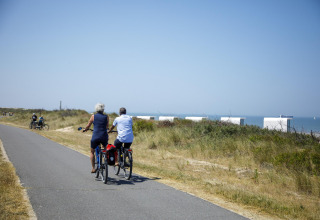 To personer cykler på en sti ved siden af strand og glamping- telte under en klar blå himmel.