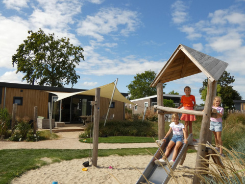 Tres niños juegan en un parque infantil junto a unas cabañas modernas de madera bajo un cielo azul.