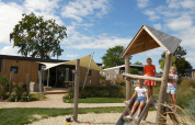 Drei Kinder spielen auf einem Spielplatz vor modernen Lodges mit Holzfassade unter blauem Himmel.