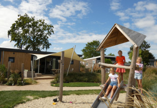 Tres niños juegan en un parque infantil junto a unas cabañas modernas de madera bajo un cielo azul.
