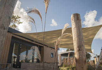 Buitenaanzicht van Dune Lodge de Luxe bij Camping Zonneweelde in Nederland, met houten gevel en schaduwdoek.