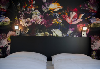 Cama con sábanas blancas y cabecero negro ante mural floral en Dune Lodge de Luxe, Camping Zonneweelde.