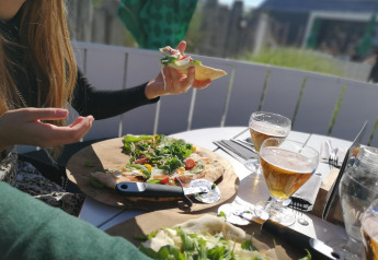 Due persone gustano pizza e birra all'aperto in un lodge, con sole e atmosfera rilassata al tavolo.