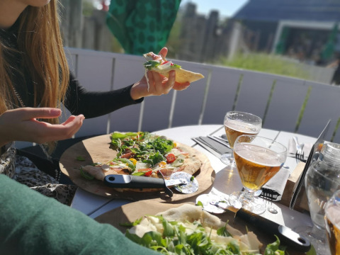 Twee mensen delen pizza en bier buiten bij een lodge, genietend van zonlicht en gezellige sfeer.