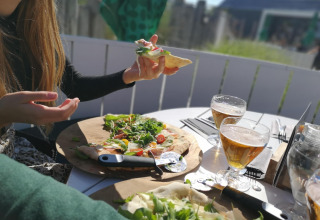 Dos personas disfrutan pizza y cerveza al aire libre en una cabaña, con sol y ambiente relajado en la mesa.