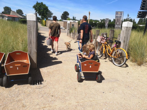 Dos hombres caminan con un niño y un perro cerca de una cabaña, con bicicletas y carritos al lado.