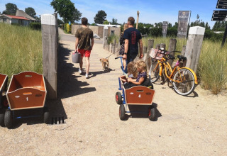 Twee mannen wandelen met een kind en een hond bij een lodge, met fietsen en karren langs het pad.