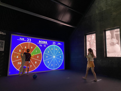 Deux personnes jouent à un jeu de points sur un écran interactif dans un lodge moderne à thème sportif.