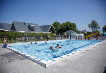 Piscina exterior en una casa de campo con niños, adultos y zona de juegos en un día soleado de verano.